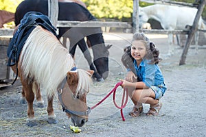 A small smiling girl with curly hair dressed in jeans feeding a pony at the stable