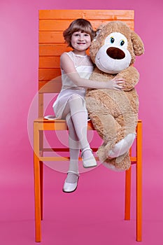 A small smiling girl sits on a huge chair and hugs a large stuffed toy bear