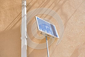 A small solar panel mounted on a metal pole is positioned next to a yellow building wall