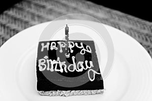 A small square chocolate birthday cake with the inscription happy birthday and one candle on a white plate. Monochrome, black and