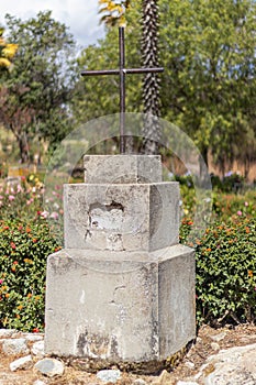 Small statue of a cross made of cement and iron, surrounded by various vegetation such as flowers in the Campo Santo