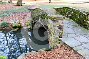 A small stone bridge covered in algae over a lake with lush green and autumn colored trees reflecting off the lake water