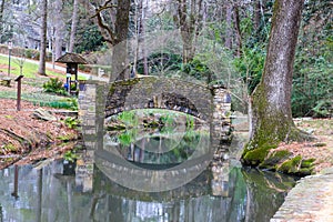 A small stone bridge covered in algae over a lake with lush green and autumn colored trees reflecting off the lake water