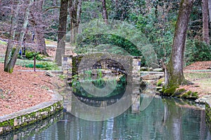 A small stone bridge covered in algae over a lake with lush green and autumn colored trees reflecting off the lake water