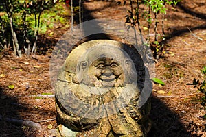 A small stone buddha statue in a Japanese garden surrounded by brown fallen pine needles and lush green trees and plants