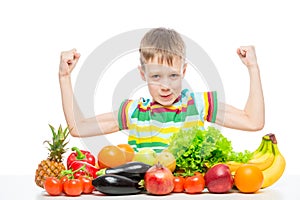 small strongman shows biceps at the table with a pile of fresh vegetables and fruits isolated