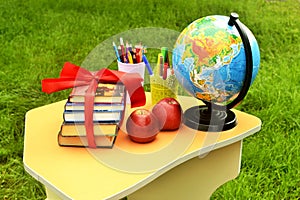 On a small student`s table against a background of green grass, there are books tied with a red gift ribbon next to a globe, multi
