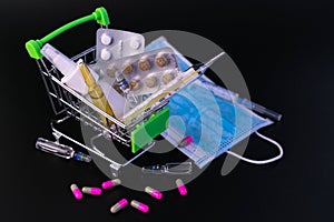 Small supermarket cart with pills, ampoules, thermometer, medical mask on a black background. Concept of protection against viral