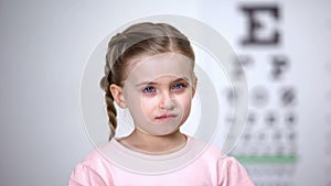 Small tearful girl looking into camera, afraid to wear glasses vision correction