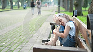 Small tired child girl sitting on a bench with closed eyes resting in summer park.