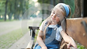 Small tired child girl sitting on a bench with closed eyes resting in summer park