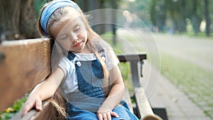 Small tired child girl sitting on a bench with closed eyes resting in summer park