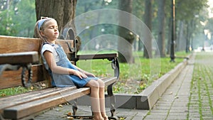 Small tired child girl sitting on a bench with closed eyes resting in summer park