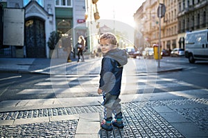 A small toddler boy standing by a road outdoors in city at sunset, looking back.