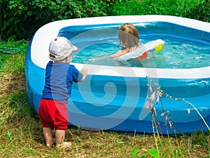 small toddler boy watches with interest at the side of the pool for his sister swimming in the water. T