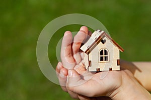 Small toy house in hands of child on green grass background. Concept mortgage, dream house, real estate acquisition.