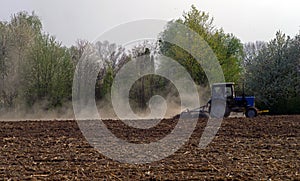 A small tractor cultivates a plowed field in the forest. Dust trail behind the tractor.