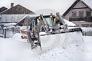 Small tractor with plough removing snow loads during heavy snowstorm calamity, village houses in background