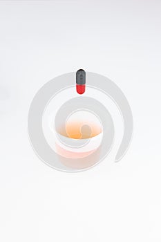 Small transparent plastic medicine drinking cup with red and black medicament capsule above it isolated on white