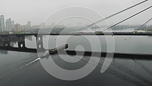 Small tugboat carrying large barge loaded with sand along wide river under concrete bridge in fog at industrial city. Scow floatin