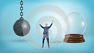 A small victorious businessman stands near a broken glass sphere and an iron wrecking ball.