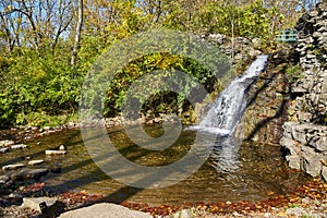 Small waterfall over rocks going into pond with green trees and walking bridge