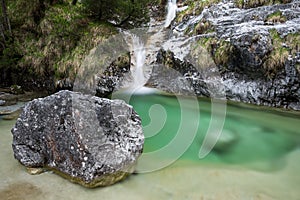 A small waterfall running into an emeral pond, with a boulder in the foreground