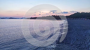 A small wave rolls onto the sandy and pebbly beach of the seashore. The sun has set behind the trees and painted the sky with clou