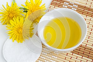 Small white bowl with cosmetic cleansing oil, dandelion flowers and cotton pad. Natural spa and beauty treatment recipe.
