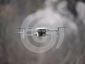 Small white drone with a built-in camera in mid-flight and a blurred background of trees