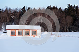 Small white hen house with orange trimmed window in snowy field seen during a beautiful blue early winter morning, St. Augustin de
