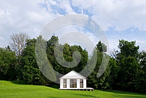 A small white house with curtained windows stands in the center of a small slope against the background of the forest