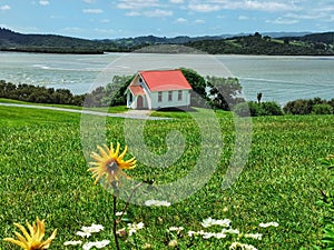 A small white wooden church building on an estuary shove with a green lawn and flowers on the foreground on the photo