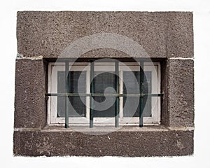 Small white wooden window with glass panes covered by steel bars in a large grey stone frame on a white wall