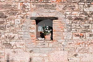 A small window under a wooden lintel decorated with a vase with white and violet flowers in a stone building of a medieval village