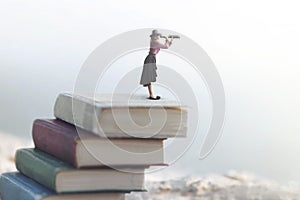 Miniature woman looks at the infinity with the spyglass on a scale of books