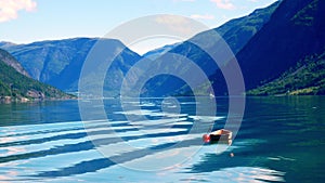 Small wooden boat moored to a red buoy bobs gently in waters of the Lustrafjord in Norway, with reflections of mountains, blue sky