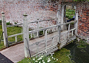 Small wooden bridge with an interesting open pattern crosses a stream and leads into a walled garden