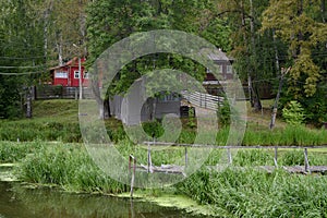 A small wooden bridge over a swampy river backwater with wooden houses on the shore in the forest on a summer evening