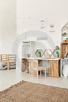 Small wooden chair standing by the desk and crate shelves in white Scandi baby girl room interior with carpet and posters on