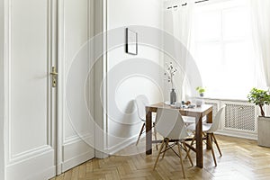 A small wooden dining table with chairs in a sunny room interior with white walls, double door and a window. Real photo.