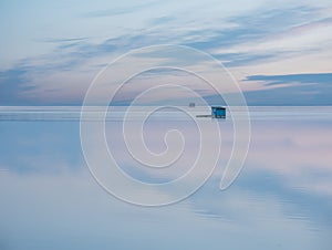 A small wooden house on the water`s edge with the reflection of the sky in the water.