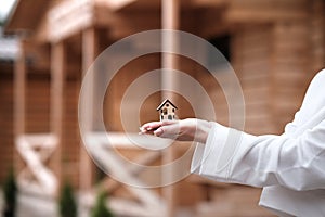 A small wooden house on a woman s hand against the background of a built wooden house. Concept of buying an ecological