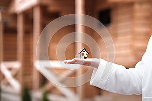 A small wooden house on a woman s hand against the background of a built wooden house. Concept of buying an ecological