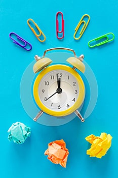 Small yellow alarm clock on blue background, Colorful office staples and crumpled paper balls. Top View. The concept of office wor
