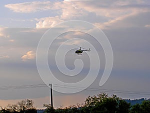 A small yellow helicopter flying over trees and power line pylons, with a blue sky in the background