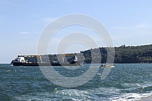 Small yellow pilot ship leads a large black cargo container ship to the seaport on a sunny day during small storm. Industrial