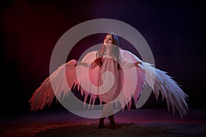 Small young model girl with white wings posing and dancing in dark black studo during photoshoot with flour or dust and colour