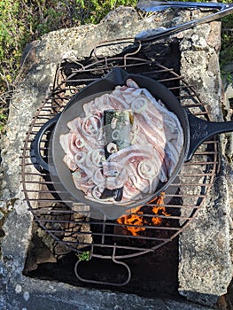 A smallmouth bass in a cast iron pan with bacon and onion slices cooking over a fire