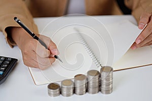 Smart and active asian businesswoman hands writing something on notebook with saving growth on stacking coin on table, meaning of